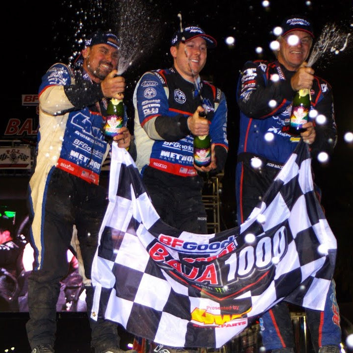 Three racers in racing suits celebrating with champagne and a checkered flag at night.